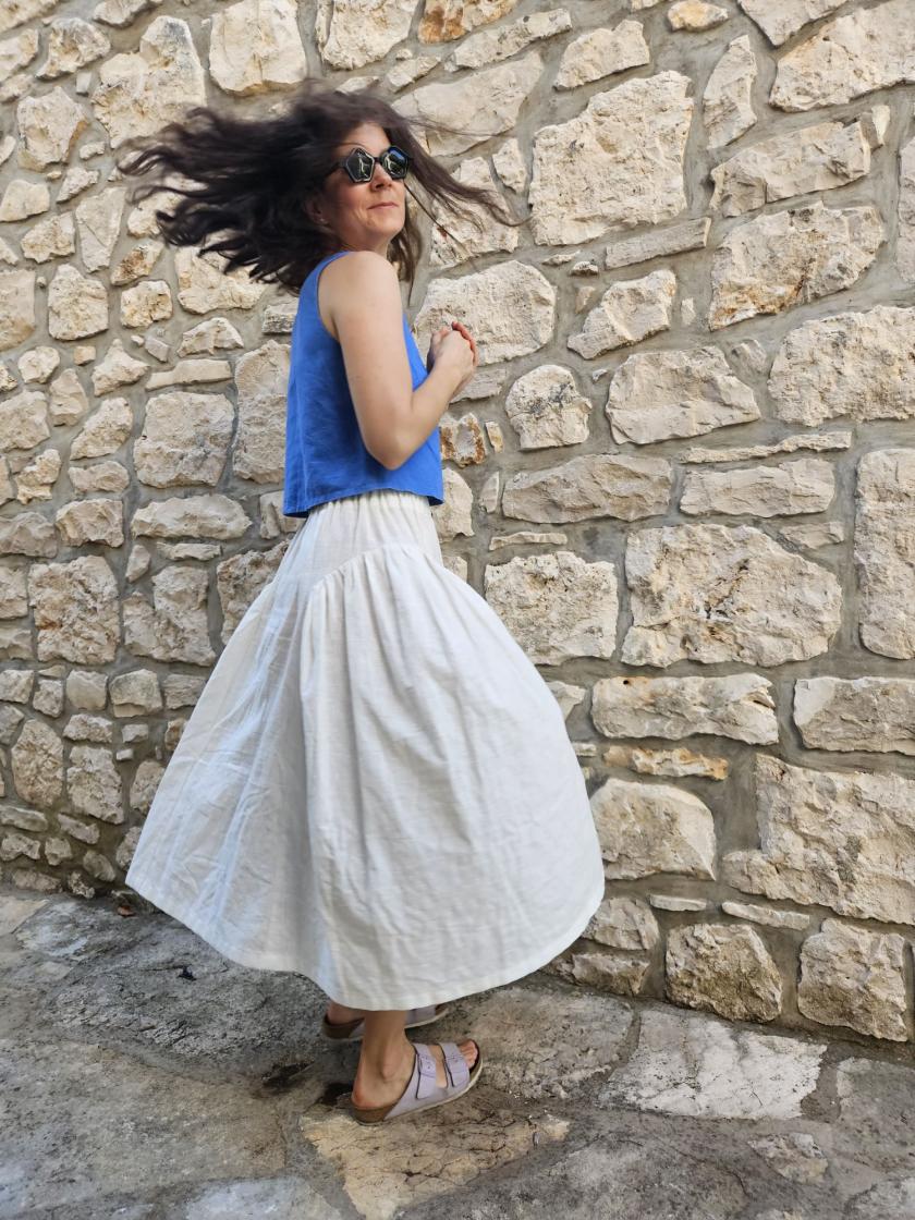 A woman is twirling in a bright blue sleeveless top and white Henrietta skirt, a sewing pattern by Spaghetti Western Sewing in front of a stone wall