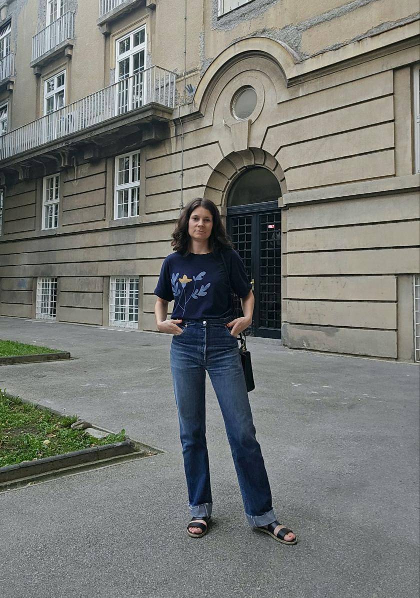 A woman is wearing blue t-shirt and refitted blue jeans made from old men's Levis using Helene jeans pattern. She is standing in front of a building looking straight, with both hands in front jeans pockets.