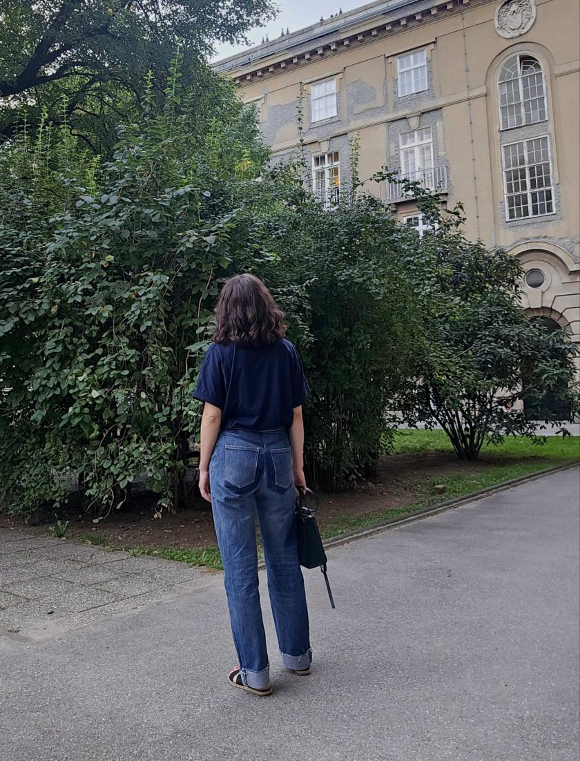 A woman is wearing blue t-shirt and refitted blue jeans made from old men's Levis using Helene jeans pattern. She is standing with her back turned to camera holding a bag in one hand in front of a building.