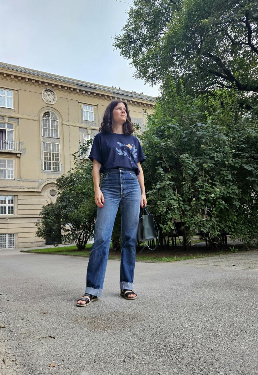 A woman is wearing blue t-shirt and refitted blue jeans made from old men's Levis using Helene jeans pattern. She is standing looking straight, holding a bag in one hand in front of a building.
